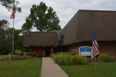 This photo shows the front door of the Library building, flanked by American flags, as seen from the parking lot.